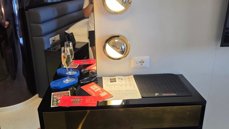 Virgin Voyages cruise ship cabin with a modern vanity, three wall lights, and reflection of bed in a round mirror.