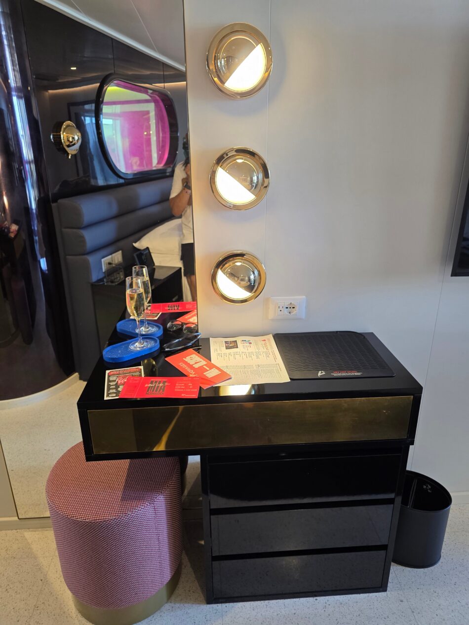 Virgin Voyages cruise ship cabin with a modern vanity, three wall lights, and reflection of bed in a round mirror.