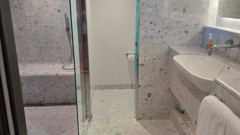 Spacious bathroom in a Virgin Voyages cruise ship cabin, featuring marble-style tiles, a glass-enclosed shower, a modern sink, and soft lighting.
