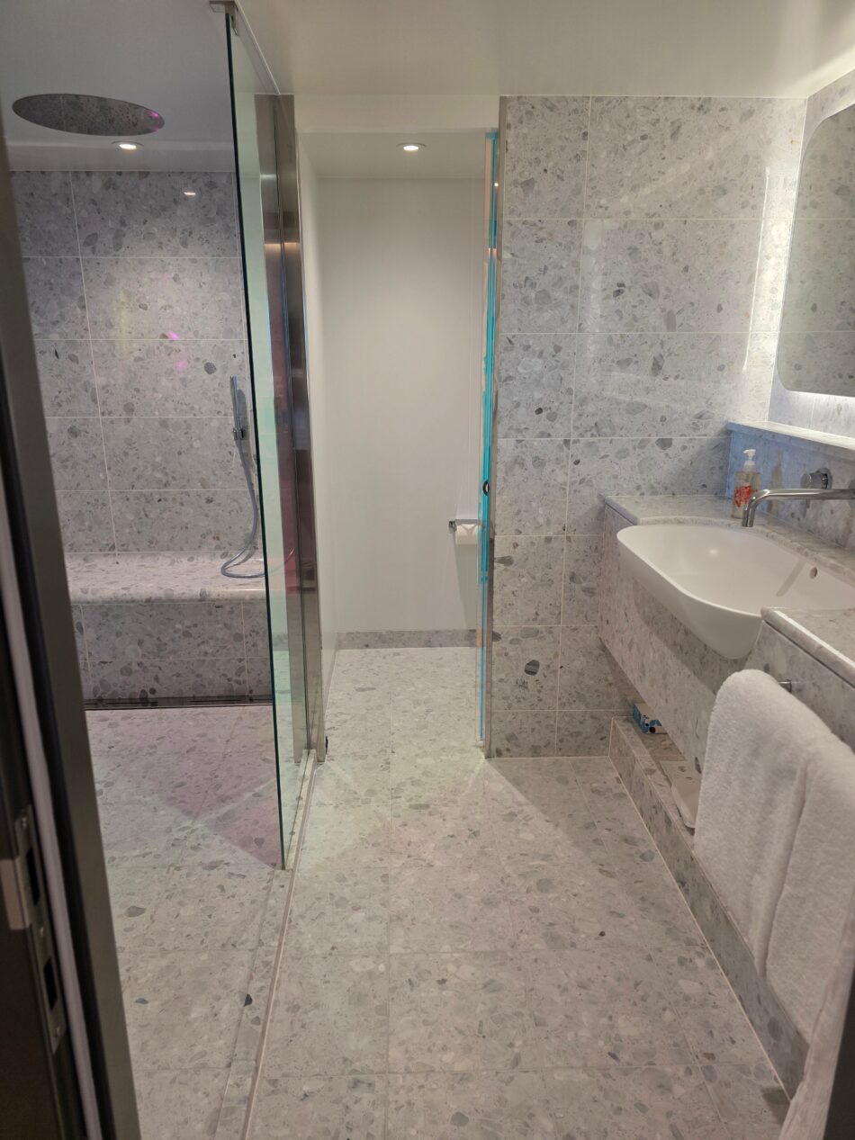 Spacious bathroom in a Virgin Voyages cruise ship cabin, featuring marble-style tiles, a glass-enclosed shower, a modern sink, and soft lighting.