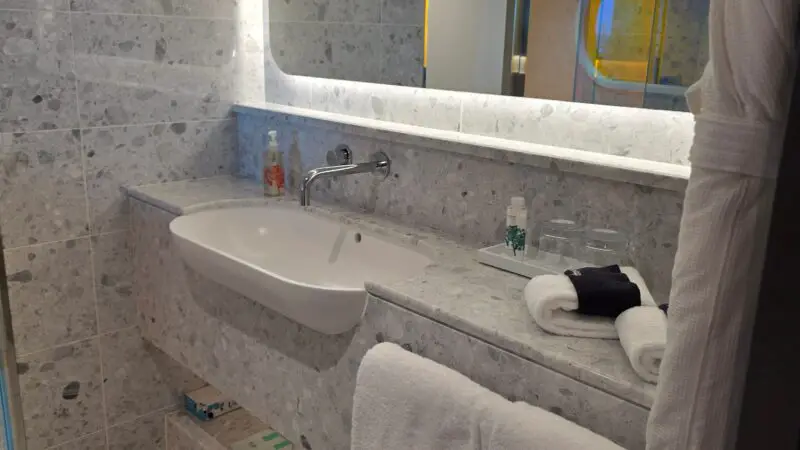 A Virgin Voyages cruise ship cabin bathroom with a modern design, featuring a large mirror, white sink, marble countertops, and neatly arranged towels.