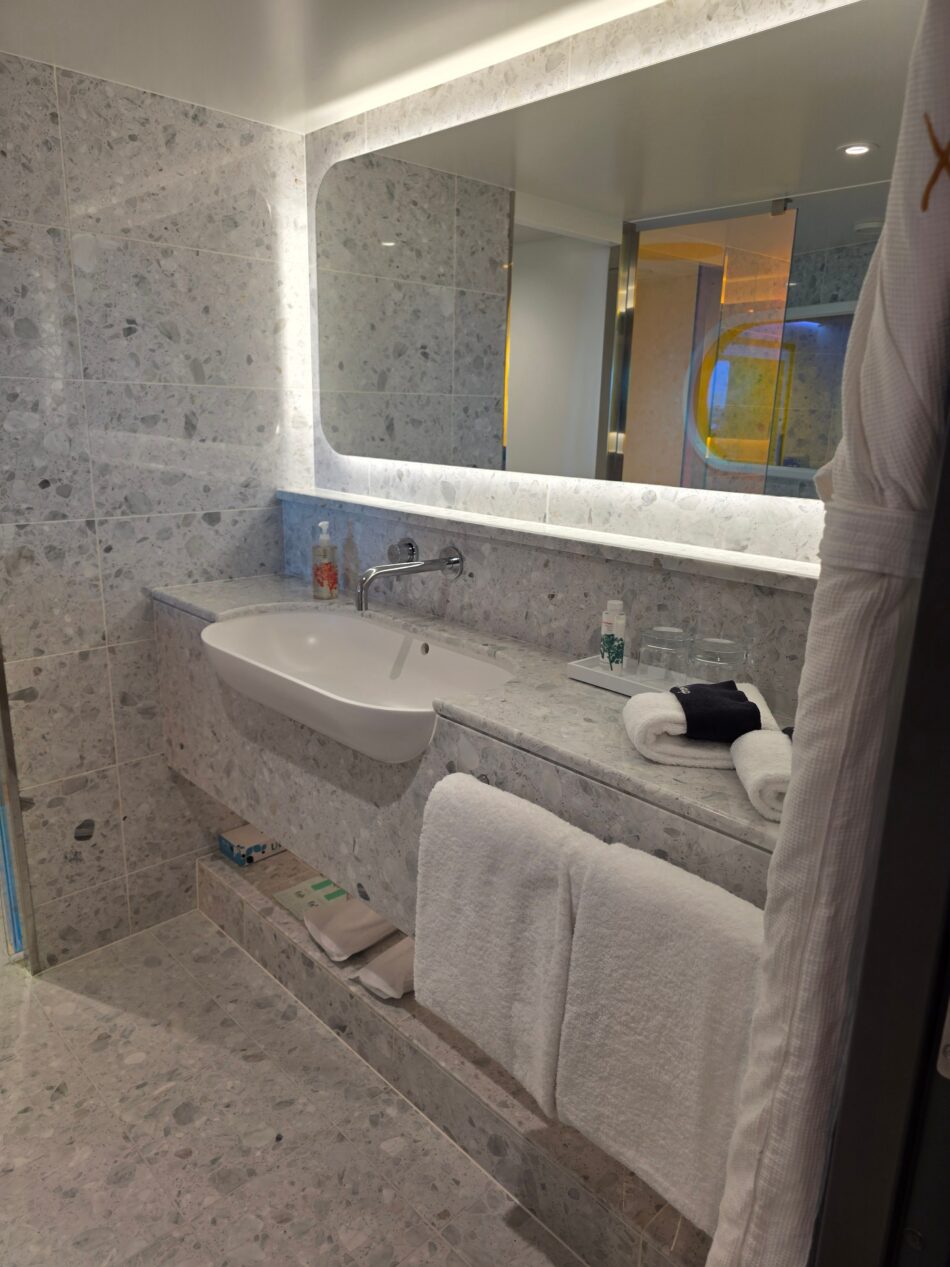 A Virgin Voyages cruise ship cabin bathroom with a modern design, featuring a large mirror, white sink, marble countertops, and neatly arranged towels.