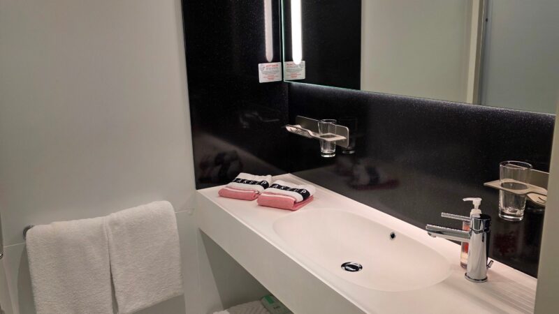 Bathroom in a Virgin Voyages cruise ship cabin, featuring a modern sink with a large mirror, towels, and toiletries.