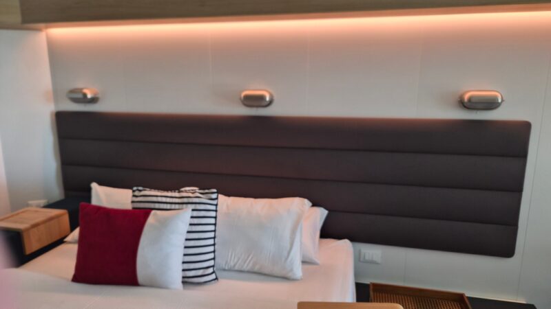 Interior of a Virgin Voyages cruise ship cabin showing a neatly made bed with a striped headboard, decorative pillows, a wooden side table, and soft ambient lighting.