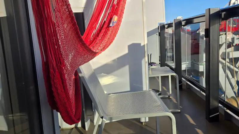 Cruise ship balcony with red hammock, two white chairs, and a small table with a beverage can.