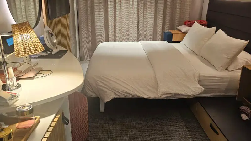 Interior of a Virgin Voyages cruise ship cabin with a bed, curtains, a round mirror, a lamp, and a TV on a wooden panel.
