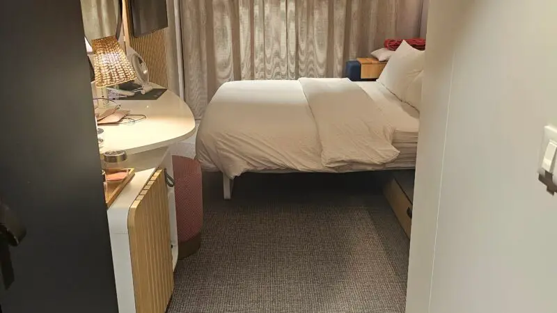 Virgin Voyages cruise ship cabin with a neatly made bed, circular mirror, TV, and a small desk area.