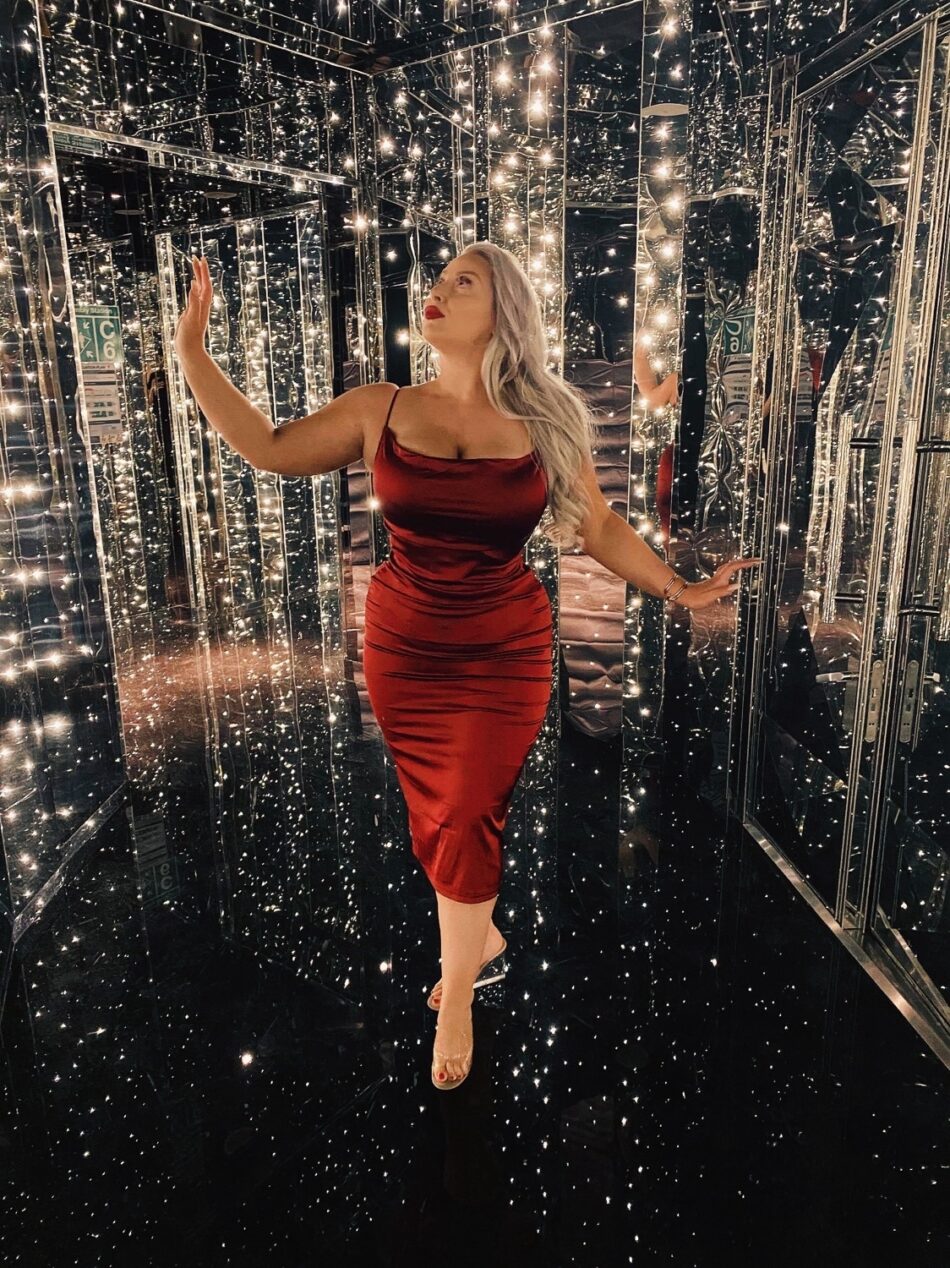 Woman in a red satin dress poses among mirrored walls with string lights on a Virgin Voyages cruise ship cabin
