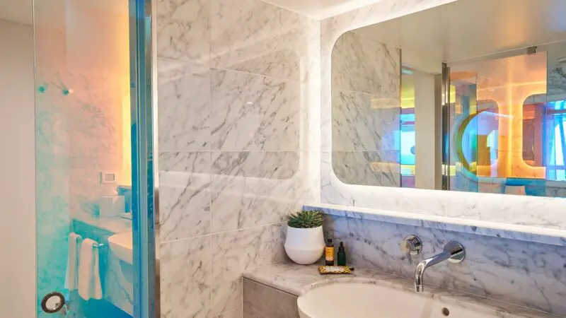 Marble bathroom with sink, large mirror, plant, and glass shower on a Virgin Voyages cruise ship cabin