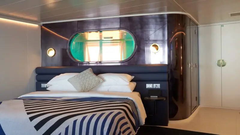 Luxurious cruise cabin with blue upholstered headboard and oval window on a Virgin Voyages ship