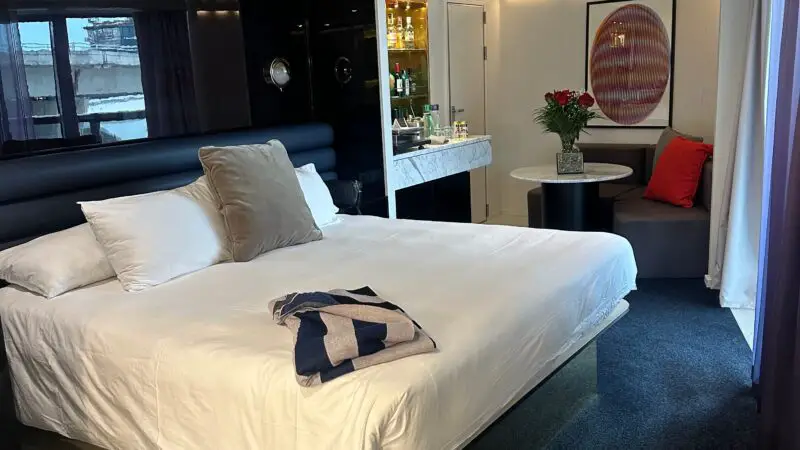 Interior of a Virgin Voyages cruise ship cabin featuring a large bed, small sitting area with a table and chairs, a minibar, and decor including a framed artwork and flowers.