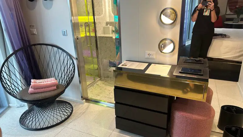 Virgin Voyages cruise ship cabin showing a modern interior with a round chair, a vanity with lights, and a bathroom entrance. A woman is reflected in the mirror.