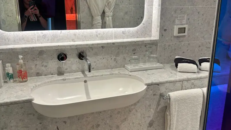 Modern bathroom in a Virgin Voyages cruise ship cabin, featuring a sleek white sink, a large backlit mirror, and marble-patterned surfaces with towels and amenities neatly arranged.