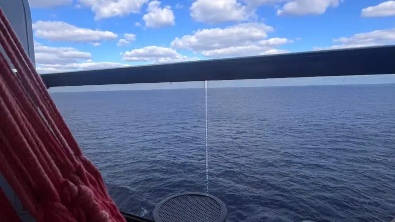 Balcony of a Virgin Voyages cruise ship cabin with a red hammock, small table, and ocean view under a partly cloudy sky.