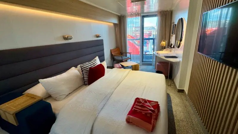 Virgin Voyages cruise ship cabin with a bed, desk, chair, and a balcony view.