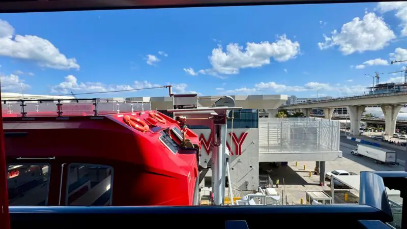 View from a Virgin Voyages cruise ship cabin balcony, showing a red ship structure with orange life rings, overlooking a sunny port area with buildings, roads, and blue sky with clouds.