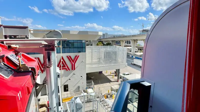 Balcony view from a Virgin Voyages cruise ship cabin, showing the Terminal V dock area with buildings and a clear blue sky.