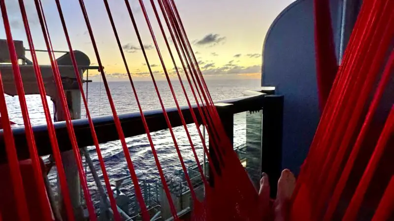 Sailor resting in a red hammock on a Virgin Voyages cruise ship balcony, overlooking the ocean at sunset.