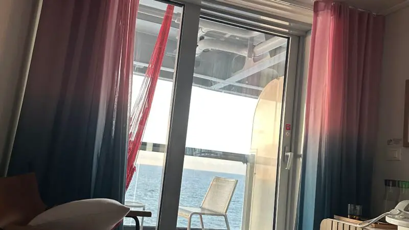 Virgin Voyages cruise ship cabin with a glass door leading to a balcony overlooking the sea, featuring colorful curtains and a lounge chair.