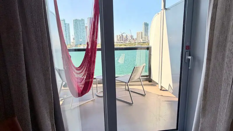 View from a Virgin Voyages cruise ship cabin showing a balcony with a red hammock, lounge chairs, and a city skyline in the background.