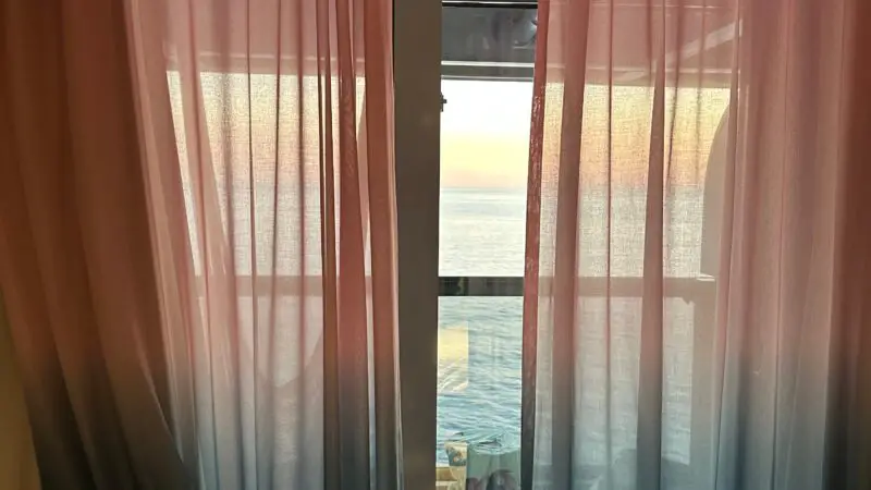 A Virgin Voyages cruise ship cabin with gradient curtains partially open, revealing a balcony view of the ocean and sunset.