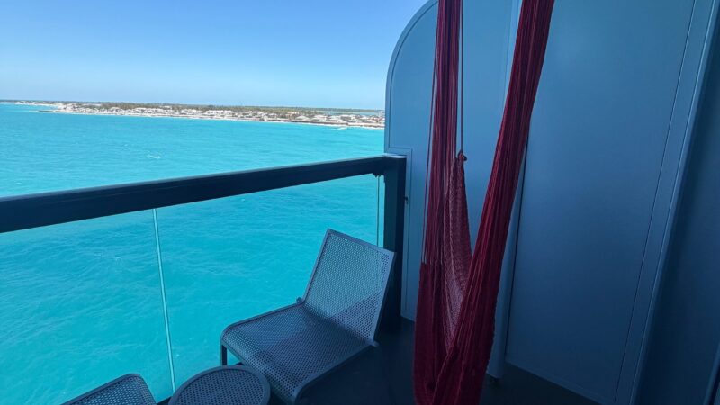 Balcony of a Virgin Voyages cruise ship cabin, featuring two chairs, a small round table, and a red hammock, overlooking clear blue ocean waters.