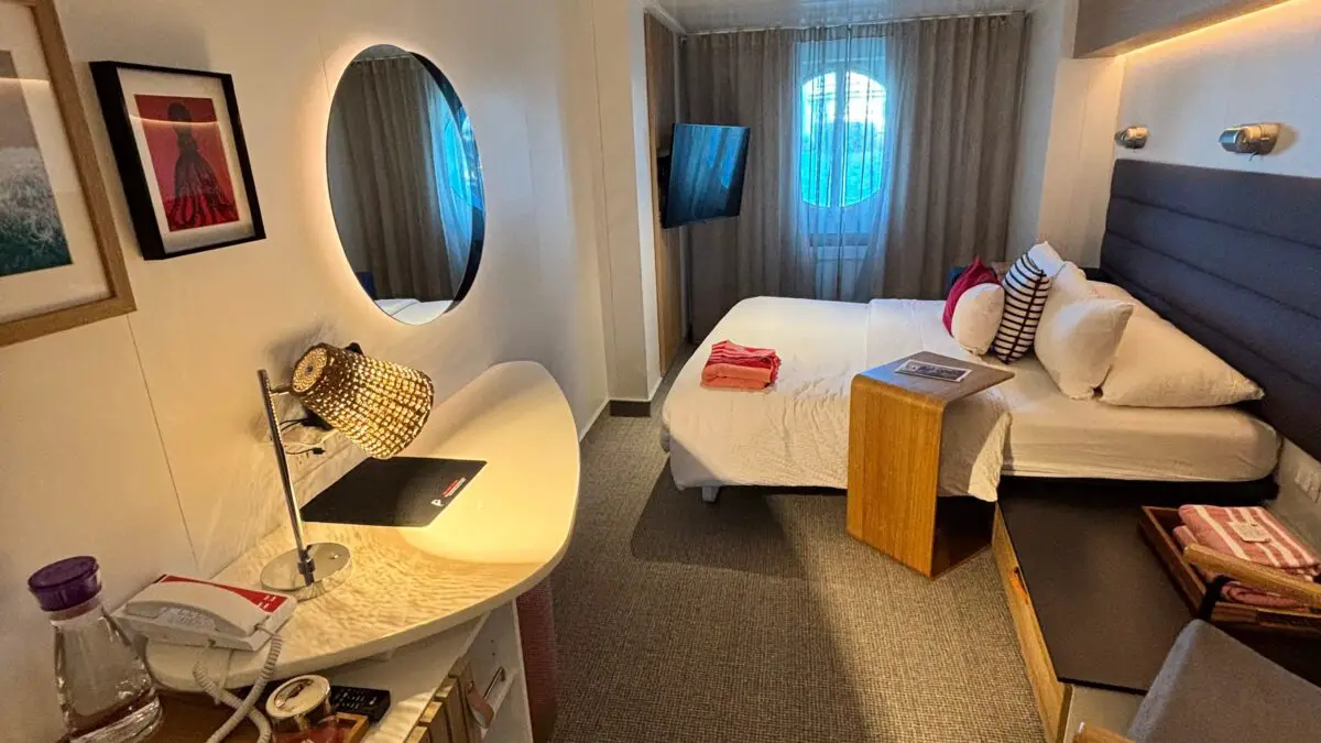 Virgin Voyages cruise ship cabin featuring a bed, wall-mounted TV, seating area, desk, and a round mirror; modern decor with soft lighting.