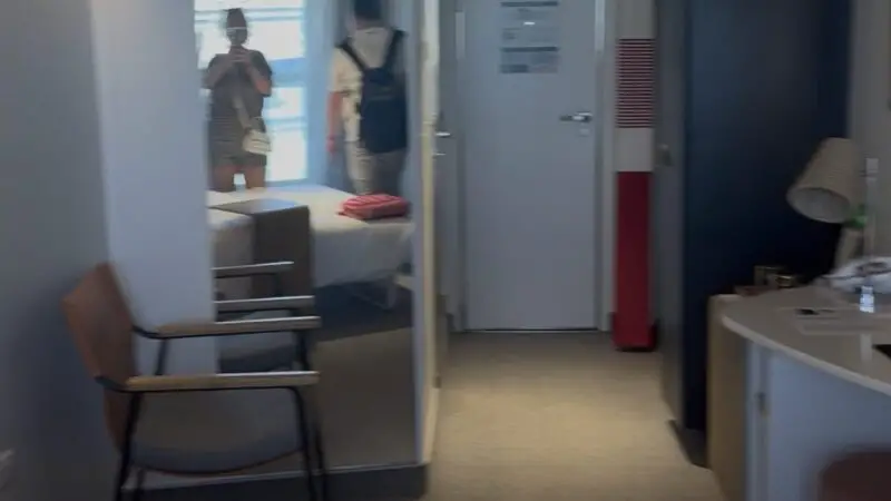 Virgin Voyages cruise ship cabin with a bed, wooden chair, desk, and mirror reflecting two people. Light spills from a window, enhancing the room's modern look.