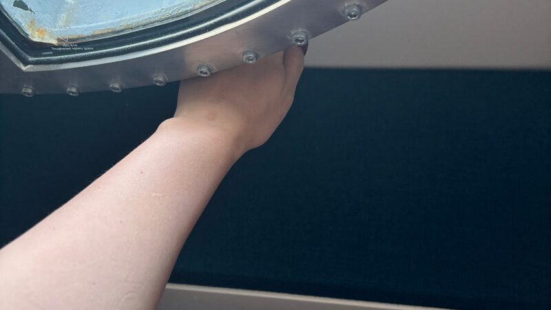 A hand reaches under a porthole window in a Virgin Voyages cruise ship cabin.