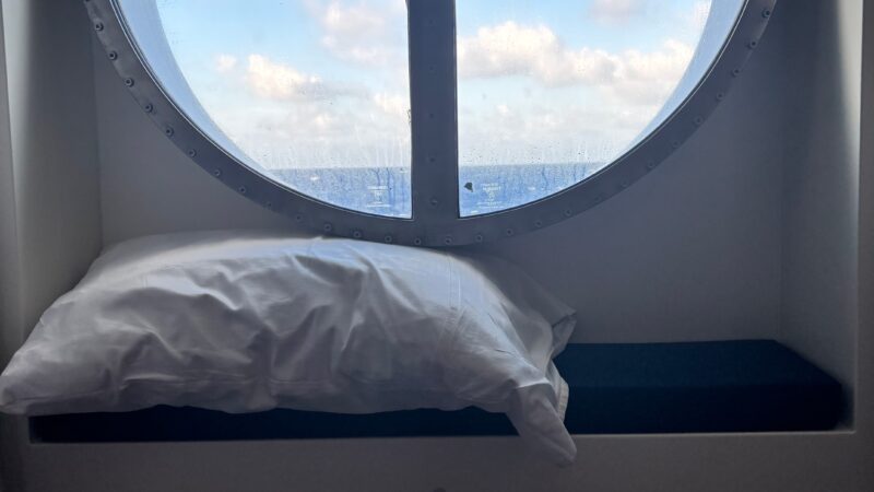 Virgin Voyages cruise ship cabin with a porthole window, showing a view of the sea and sky, and a pillow resting on the porthole ledge.
