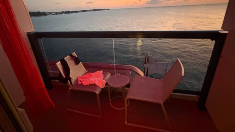 Cruise ship cabin balcony with two chairs, a small table, and a sea view at sunset.