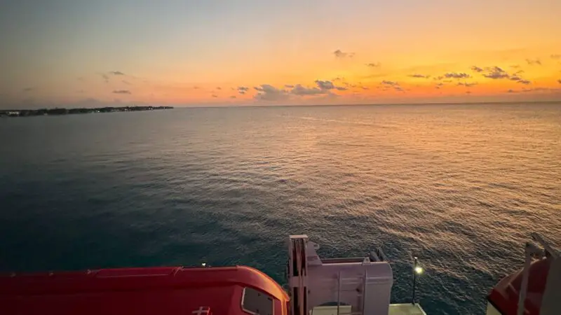 Sunset view from a Virgin Voyages cruise ship balcony, overlooking the ocean with a lifeboat below.