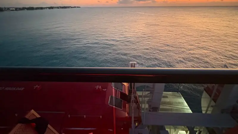 Balcony view from a Virgin Voyages cruise ship cabin at sunset, overlooking the ocean with a chair and table in the foreground.