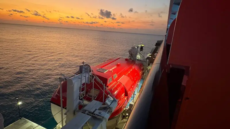 View from a Virgin Voyages cruise ship balcony overlooking the ocean at sunset, with a red lifeboat visible on the deck below.