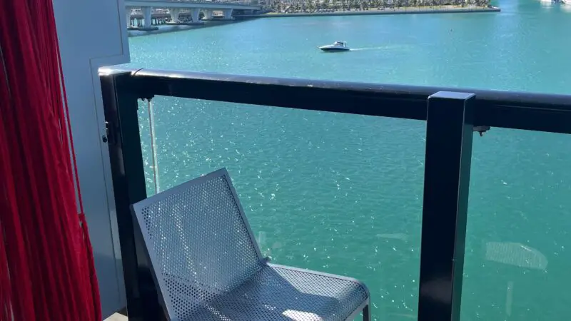 Balcony of a Virgin Voyages cruise ship cabin with metal chairs, a small table, and a view of the city skyline and water.