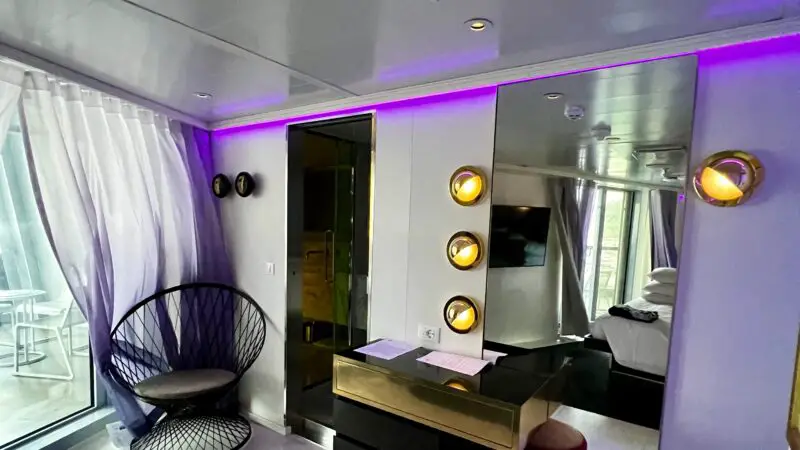 Virgin Voyages cruise ship cabin interior with modern decor, featuring a black chair, vanity desk with a mirror, colorful lighting, and a glimpse of a bed and balcony.