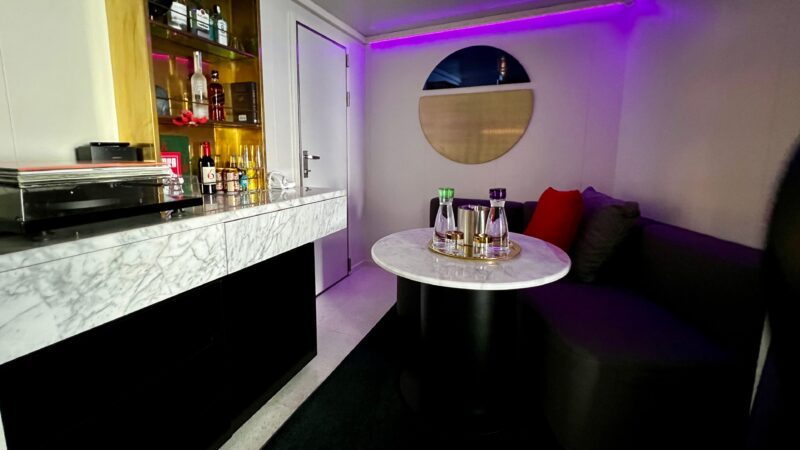 Modern cruise ship stateroom with a marble counter, stocked bar, lounge seating, round mirror, and ambient purple lighting.