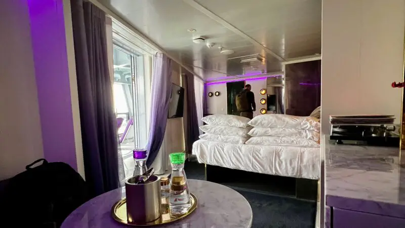 Virgin Voyages stateroom featuring a bed, marble table with drinks, and balcony access with purple lighting.
