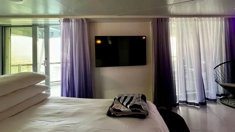 Virgin Voyages cruise ship cabin with a bed, folded towel, wall-mounted TV, floor-to-ceiling windows, and balcony access.