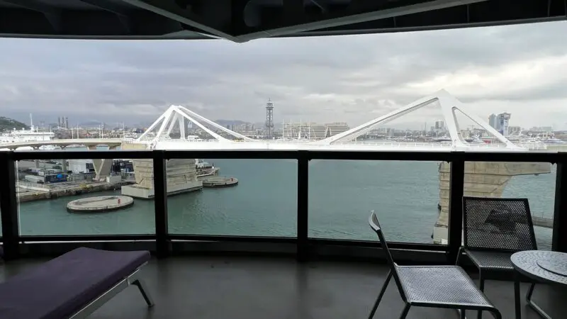 Balcony view from a Virgin Voyages cruise ship cabin overlooking a harbor with a modern white bridge and cityscape in the background.