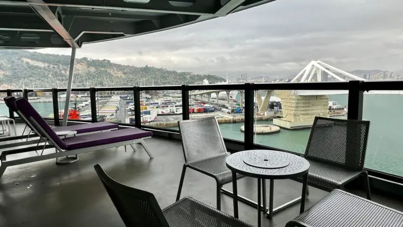 Spacious Virgin Voyages cruise ship balcony with purple lounge chairs and a view of the port and bridge.