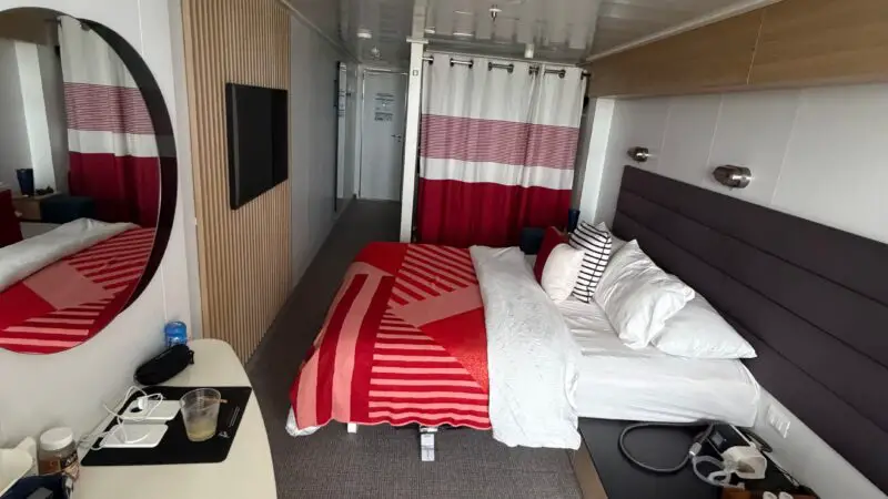 Interior view of a Virgin Voyages cruise ship cabin featuring a bed with red and white bedding, a wall-mounted TV, and a desk with various items.