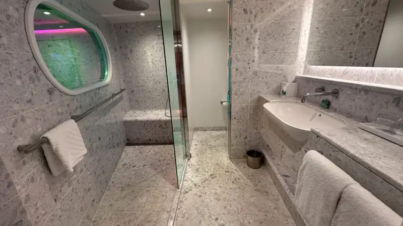 Modern Virgin Voyages cruise ship cabin bathroom with marble tiles, a glass-enclosed shower, sink, and mirrored wall.