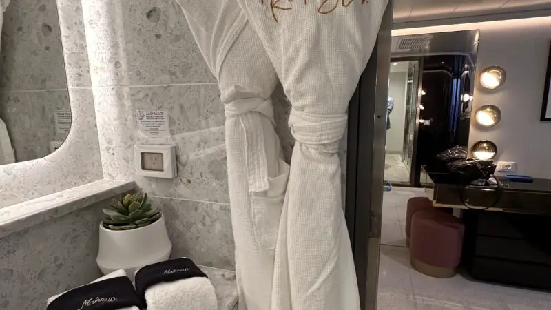 Cruise ship cabin bathroom with a hanging white robe labeled "RockStar," folded towels, and a small plant on a marble counter.