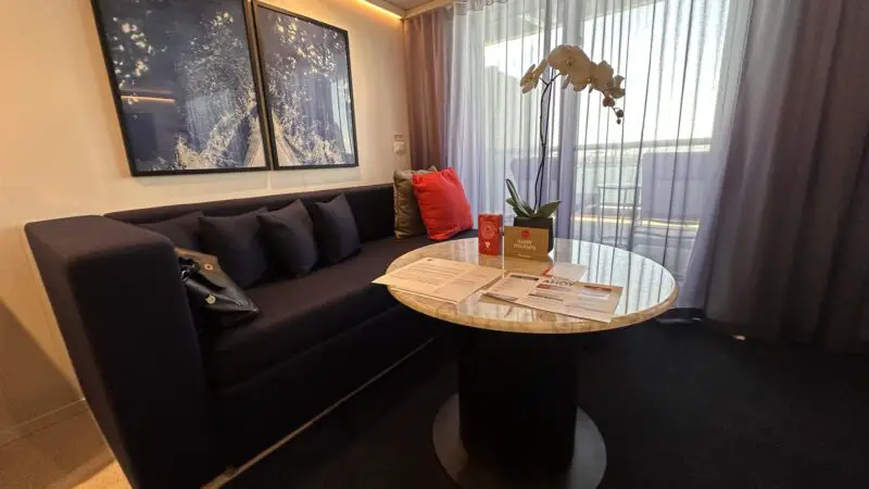 Virgin Voyages cruise ship cabin featuring a navy sofa, two artworks, a round marble table with a potted orchid and brochures, and sheer curtains leading to a balcony.