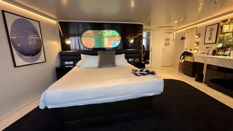 Cruise ship cabin with a large bed, circular window, modern artwork, and a bar area on the side.