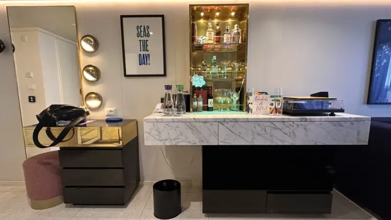 Virgin Voyages cruise ship cabin with a marble-topped bar, mirrored drawers, a small stool, and a decorative sign reading "Seas the Day!"