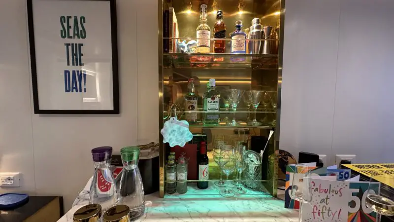 Virgin Voyages cruise ship cabin minibar with bottles, glasses, and a framed "Seas the Day!" poster.