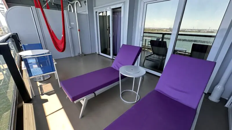 Virgin Voyages cruise ship balcony with two purple lounge chairs, a small table, a blue chair set with a round table, and a red hammock, overlooking the water.