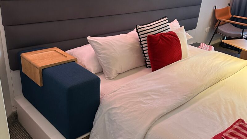 Virgin Voyages cruise ship cabin featuring a bed with white and red pillows, gray upholstered headboard, and ambient lighting. A chair and small table are visible in the background.